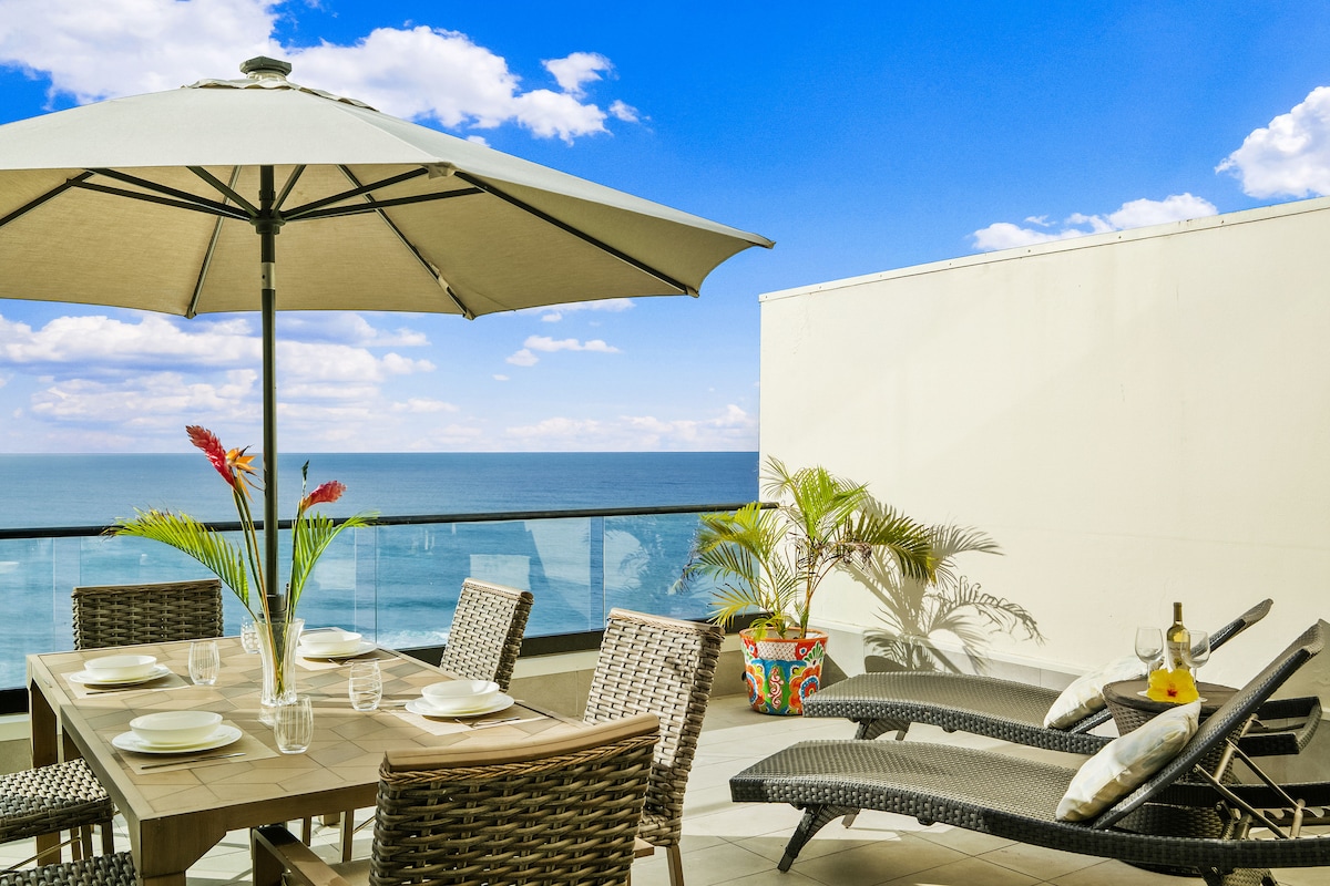 An outdoor lanai features a dining table set for six, accompanied by two chaise loungers. A large umbrella provides shade, while ocean views and blue skies enhance the serene setting. A colorful planter and tropical decor add vibrant touches to the space.