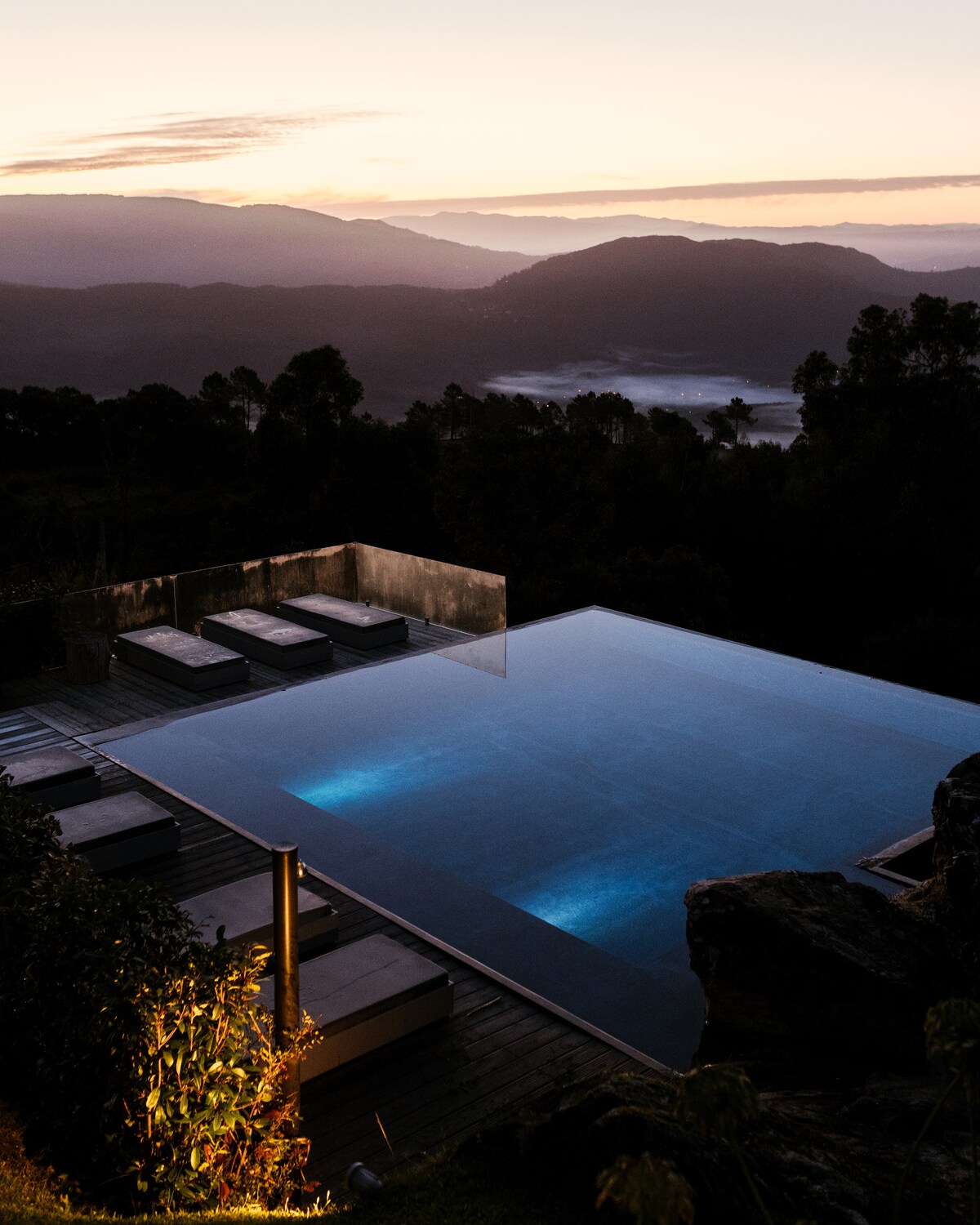 An infinity pool is seen against a backdrop of rolling mountains during sunset. The water reflects the soft hues of the sky, while lounge chairs line the pool's edge. Surrounding foliage adds a natural touch to the serene landscape.