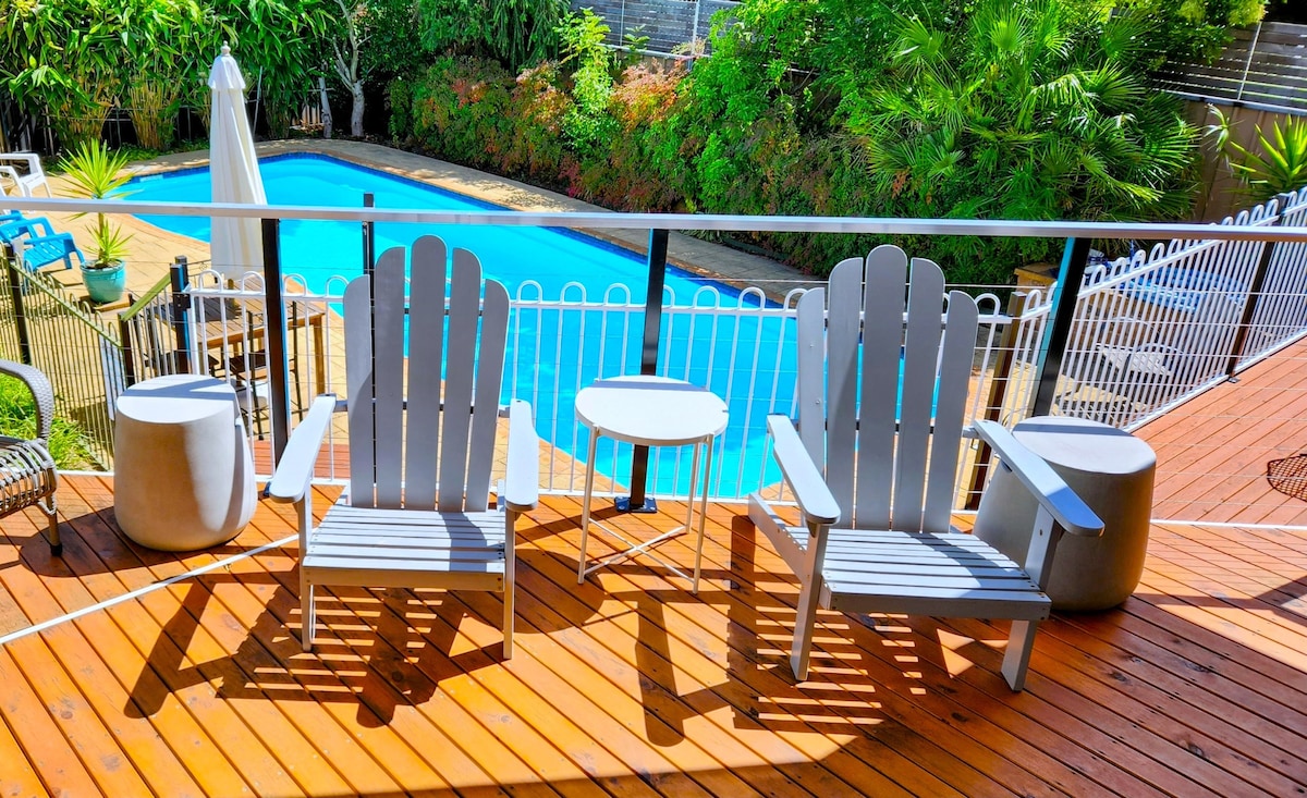 A spacious deck features two comfortable gray chairs flanking a small round table. A clear blue pool glimmers in the background, surrounded by lush greenery, offering a serene outdoor atmosphere. The area is well-lit, inviting relaxation by the water.