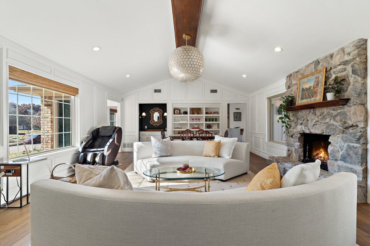 A spacious living area features a curved sectional sofa, a glass coffee table, and a cozy fireplace with stacked stone. Natural light fills the room through large windows, accented by warm wood beams overhead. Elegant decor includes a round mirror and tasteful artwork.