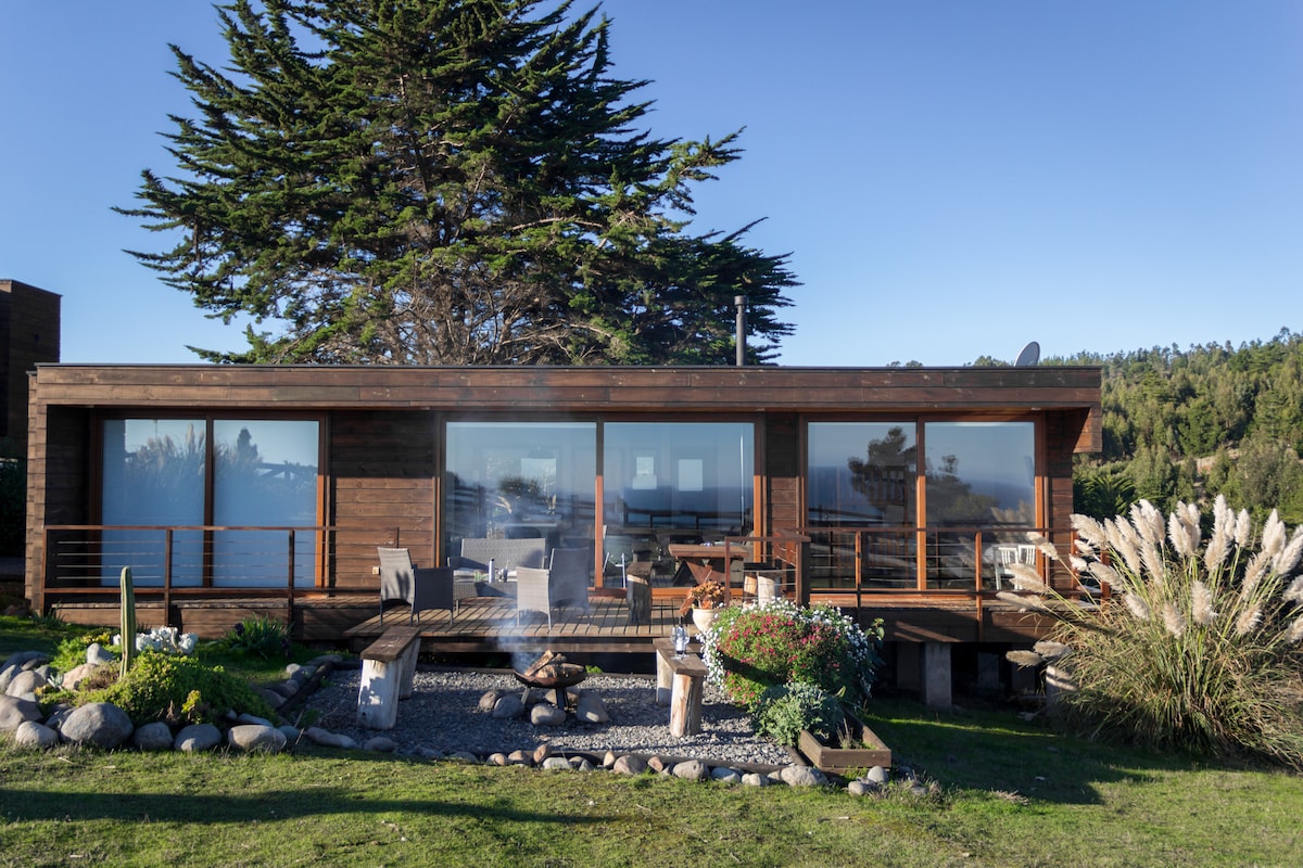 A wooden house is set amidst a natural landscape, showcasing expansive glass windows that offer views of the outdoors. A spacious deck features comfortable seating and a fire pit, surrounded by greenery and decorative stones, enhancing the connection to nature.
