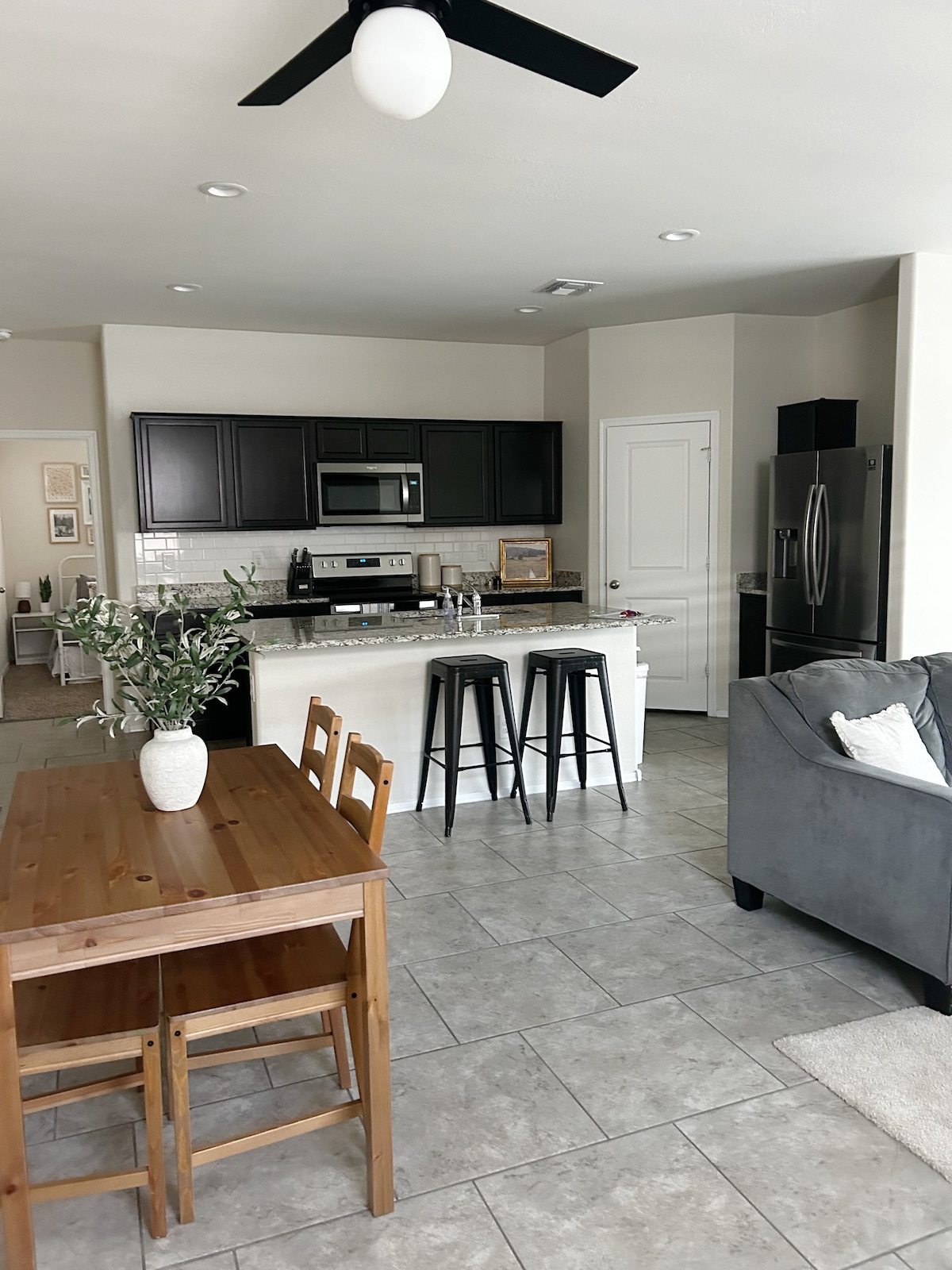 An open-concept kitchen and dining area is featured, showcasing dark cabinetry and stainless steel appliances. A wooden dining table is surrounded by four chairs, while sleek bar stools are positioned at the kitchen island. Soft gray furnishings provide comfort in the adjacent living space.