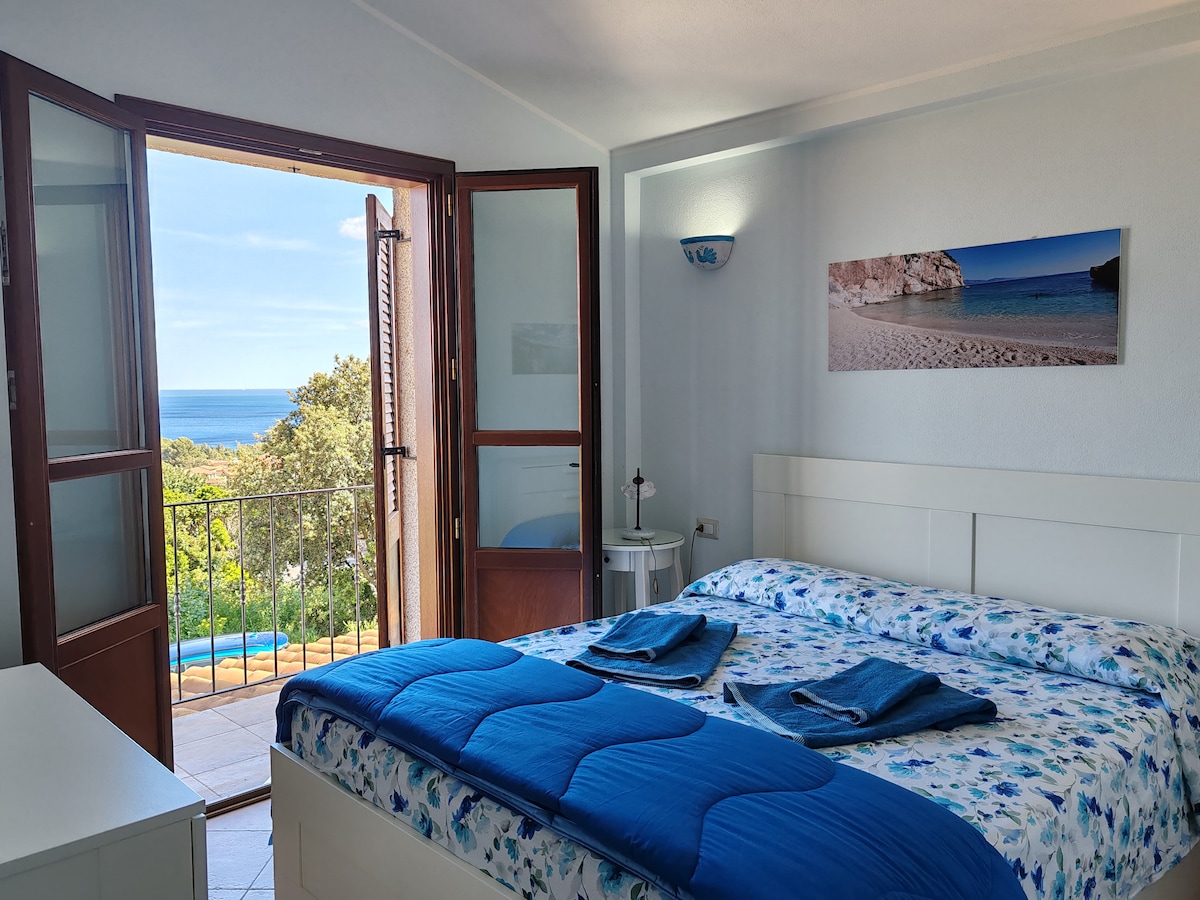 A well-lit bedroom features a spacious bed adorned with a blue blanket. Light filters through open double doors, revealing a balcony with panoramic sea views. A small nightstand sits beside the bed, and a piece of coastal artwork hangs on the wall.