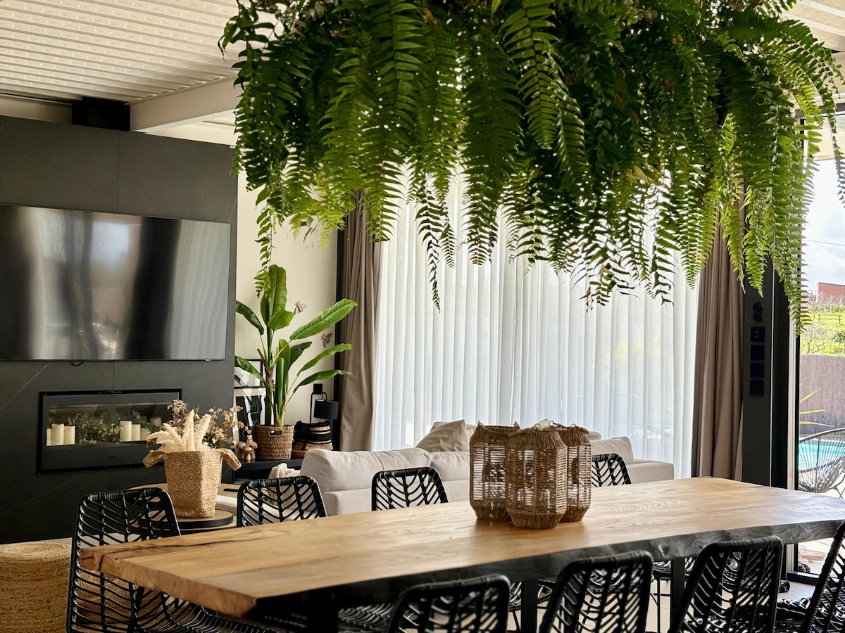 A modern dining area is presented with a large wooden table surrounded by woven black chairs. A wall-mounted television is visible, and lush green ferns hang from the ceiling. Natural light filters through sheer curtains, complementing the inviting decor and nearby plants.