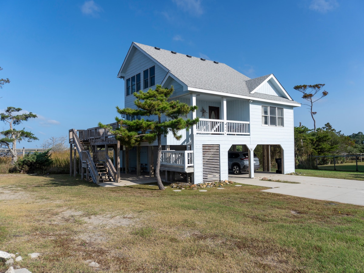 A coastal home stands on a grassy lot, featuring a light blue exterior and a gabled roof. A spacious deck is accessible via stairs, and parking is available beneath the house. Nearby, windswept trees complement the serene surrounding landscape.