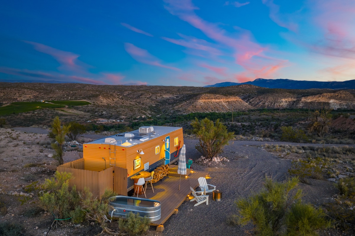 A singular camper is positioned on a gravel area, surrounded by desert vegetation and panoramic mountain views. Outdoor seating is arranged on the deck, which includes a round table and chairs. A stock tank pool is located nearby, reflecting the colors of the evening sky.