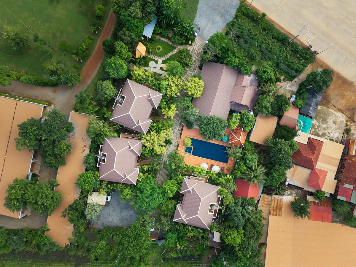 An aerial view captures a cluster of well-maintained structures surrounded by lush greenery. A central swimming pool is visible, framed by tropical plants and trees. The surrounding pathways weave through the landscape, enhancing the serene atmosphere of the property.