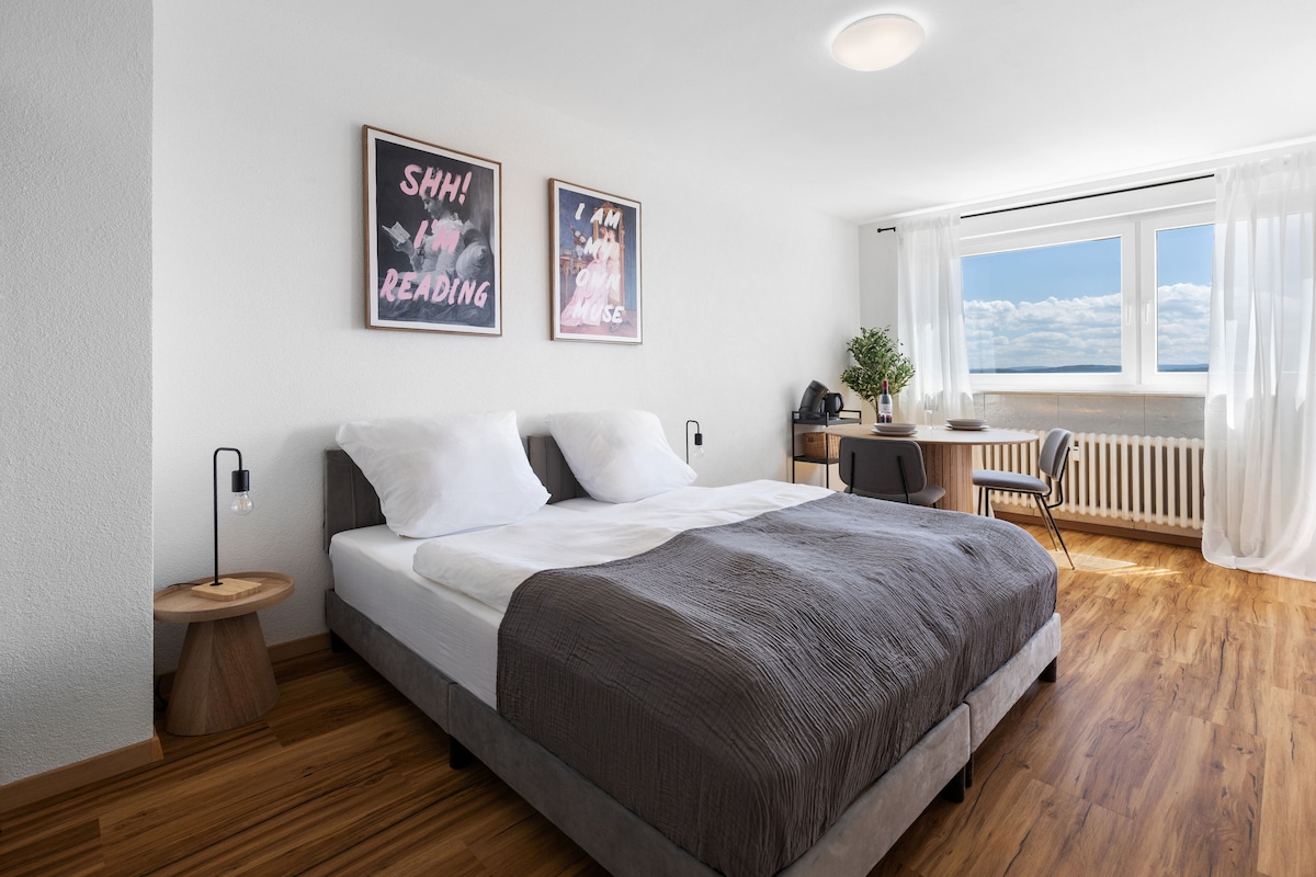 A spacious sleeping area features a large bed with a light grey coverlet and white pillows. Art prints are displayed above the bed. A small round table with a plant and a work desk are positioned near a large window, offering ample natural light.
