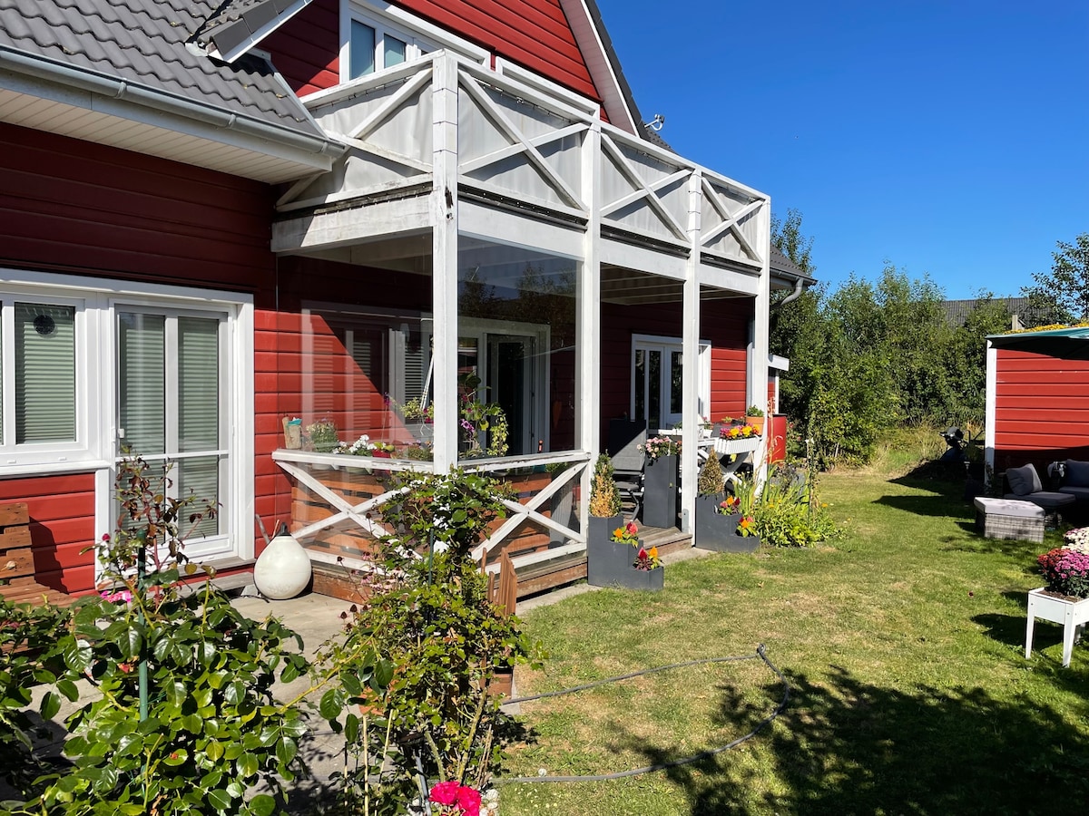 A red house with a spacious porch is surrounded by vibrant flower arrangements and green grass. Large windows allow natural light to fill the interior. The area features various potted plants, creating an inviting outdoor space for relaxation.