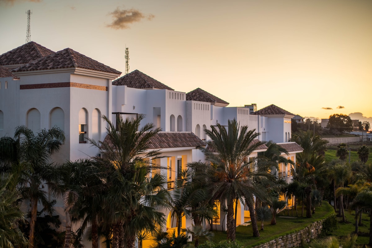 A collection of elegant white buildings is framed by lush palm trees, with soft warm light emanating from the windows at dusk. The rooftops are adorned with terracotta tiles, and a gentle gradient of sunset colors fills the sky behind the structures.