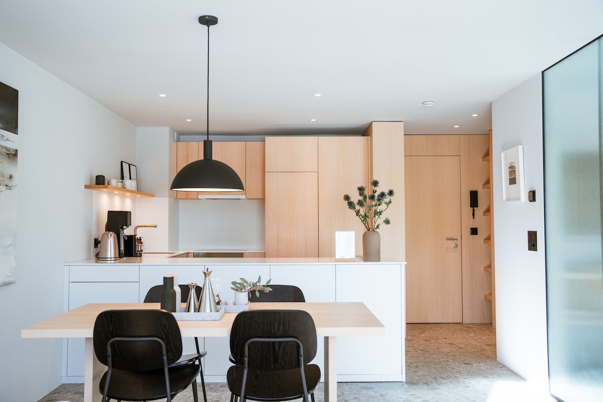 A modern kitchen and dining area showcases a light wood cabinetry, complemented by a simple table set for four. A stylish pendant light hangs above the table, while the kitchen features sleek appliances, enhancing the open and airy feel of the space.
