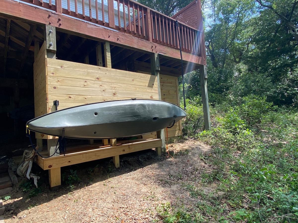 A kayak rests securely on a wooden rack beneath a cottage. The structure is surrounded by lush greenery, with sunlight filtering through the trees. The ground is covered in natural soil and leaves, providing a serene outdoor setting.