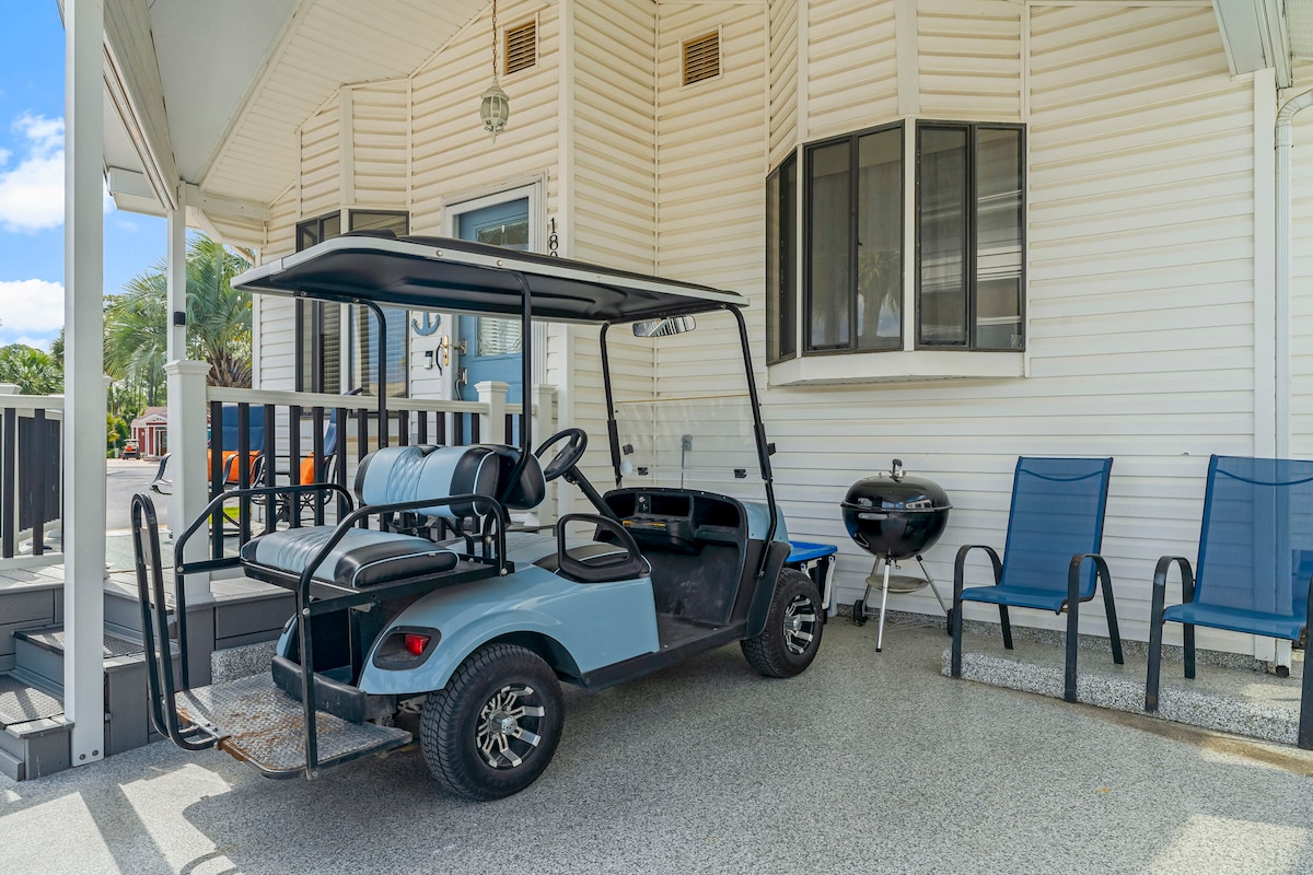 A golf cart is parked on a covered porch beside two blue outdoor chairs and a small charcoal grill. The porch features a light-colored exterior wall with a large window, allowing natural light into the area.