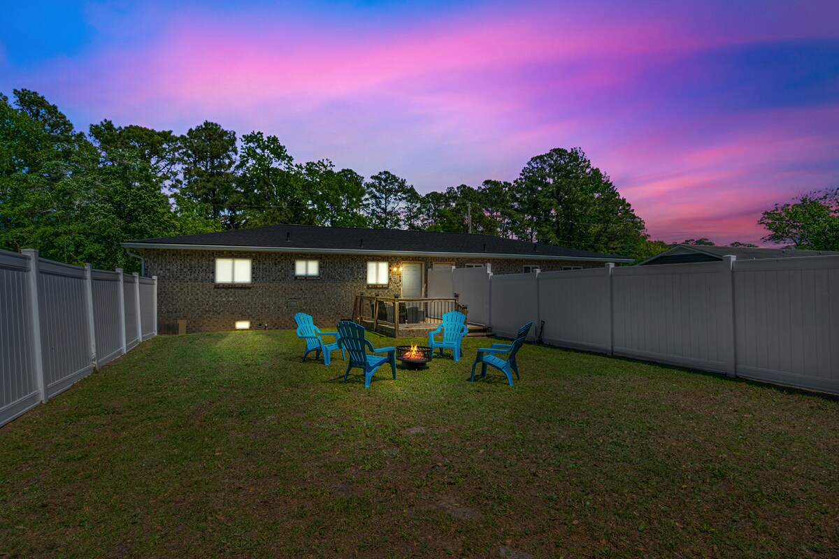 The large fenced backyard features a cozy fire pit surrounded by colorful outdoor chairs. A welcoming home can be seen in the background, contrasted against a vibrant sunset sky filled with shades of pink and purple.