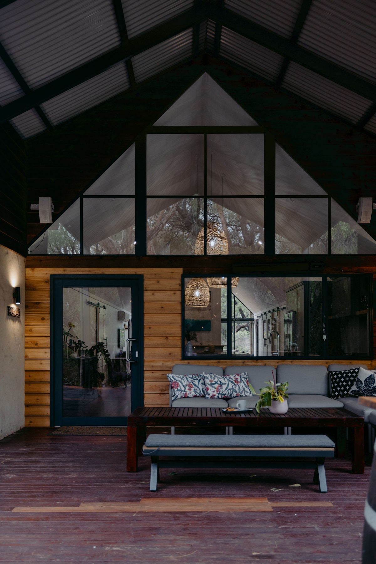 An indoor lounge area features high, angled ceilings and large glass panels that allow natural light to fill the space. A cozy seating arrangement includes patterned cushions on a wooden bench and a coffee table in front, complemented by warm wooden accents and contemporary decor.