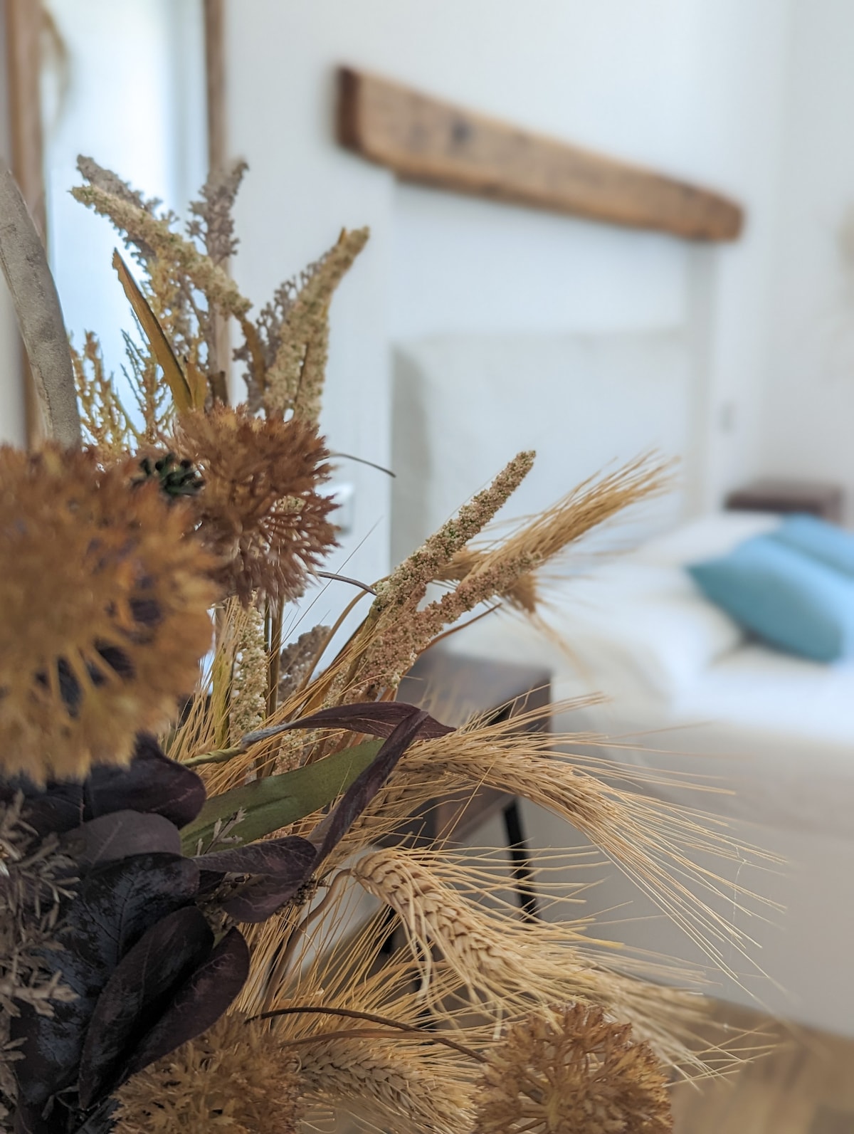 A delicate arrangement of dried flowers adds a natural touch to the room’s decor. In the background, a spacious bed with soft linens is visible, complemented by decorative cushions. Natural light illuminates the space, creating a serene and calming atmosphere.
