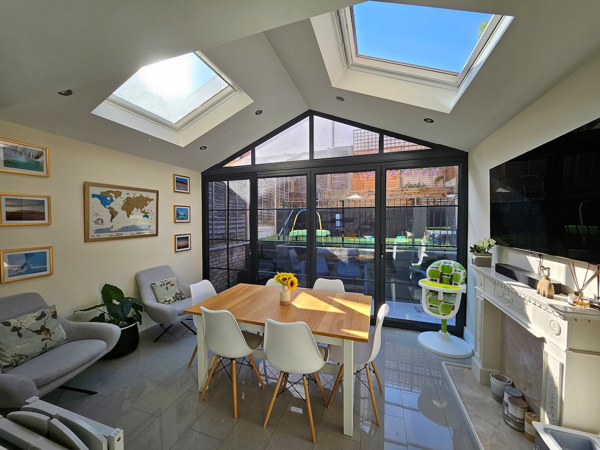 A light-filled dining area features a large wooden table surrounded by white chairs. Skylights provide ample natural light, while large glass doors open to a private garden. Neutral tones and minimal decor highlight the space, complemented by a wall-mounted TV and decorative elements.