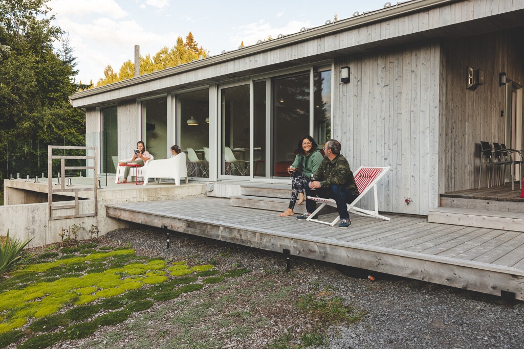 A wooden deck features a mix of seating options, including a lounge chair and two casual chairs, in a natural setting. Large glass doors reveal the inviting interior space. Lush greenery and moss enhance the outdoor experience, creating a serene atmosphere.