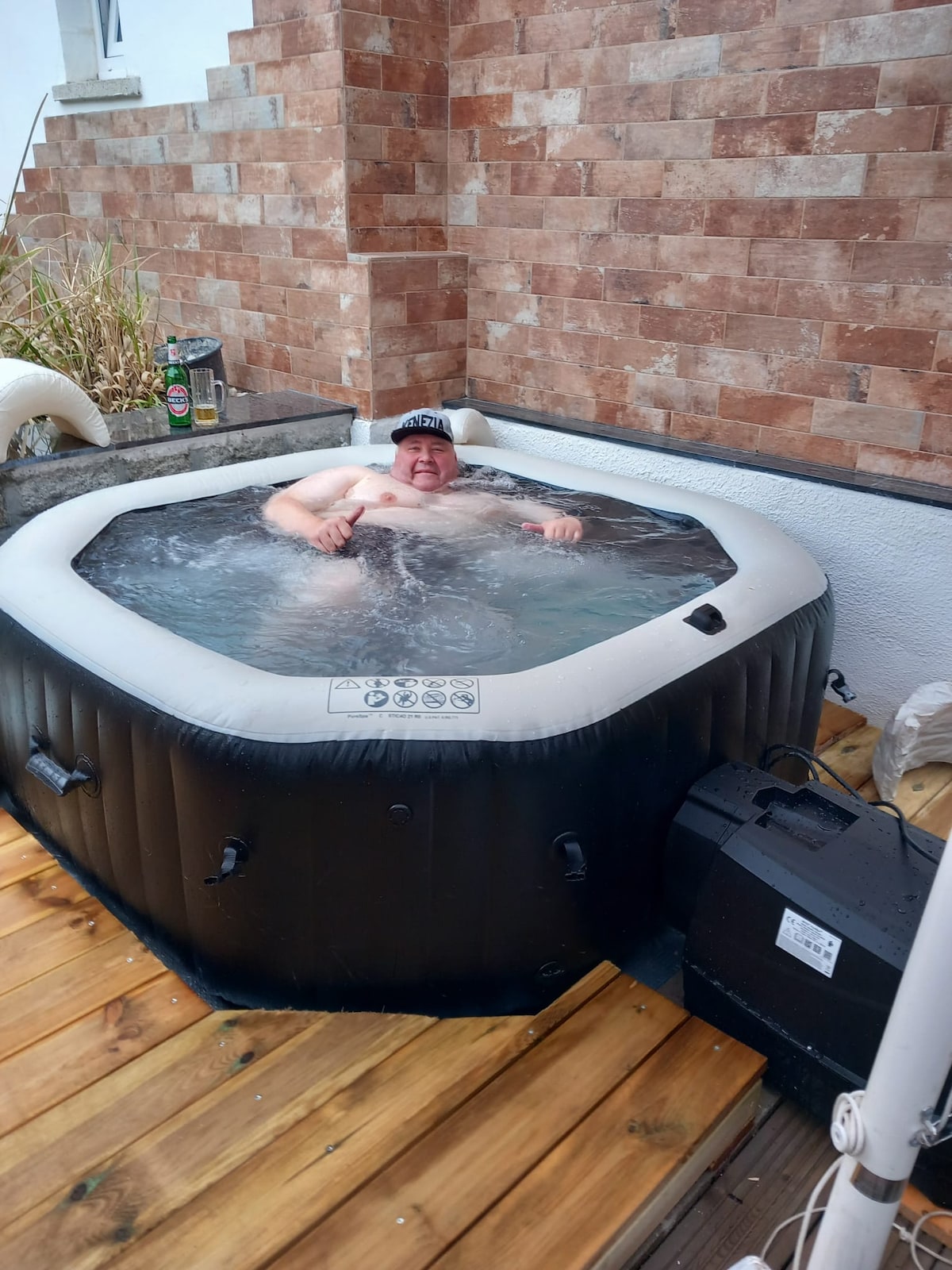 A private hot tub is situated on a wooden deck, surrounded by brick walls. Bubbles can be seen in the water, inviting relaxation. The tub is equipped with a control unit placed nearby. Lush greenery is partially visible in the background, enhancing the serene environment.