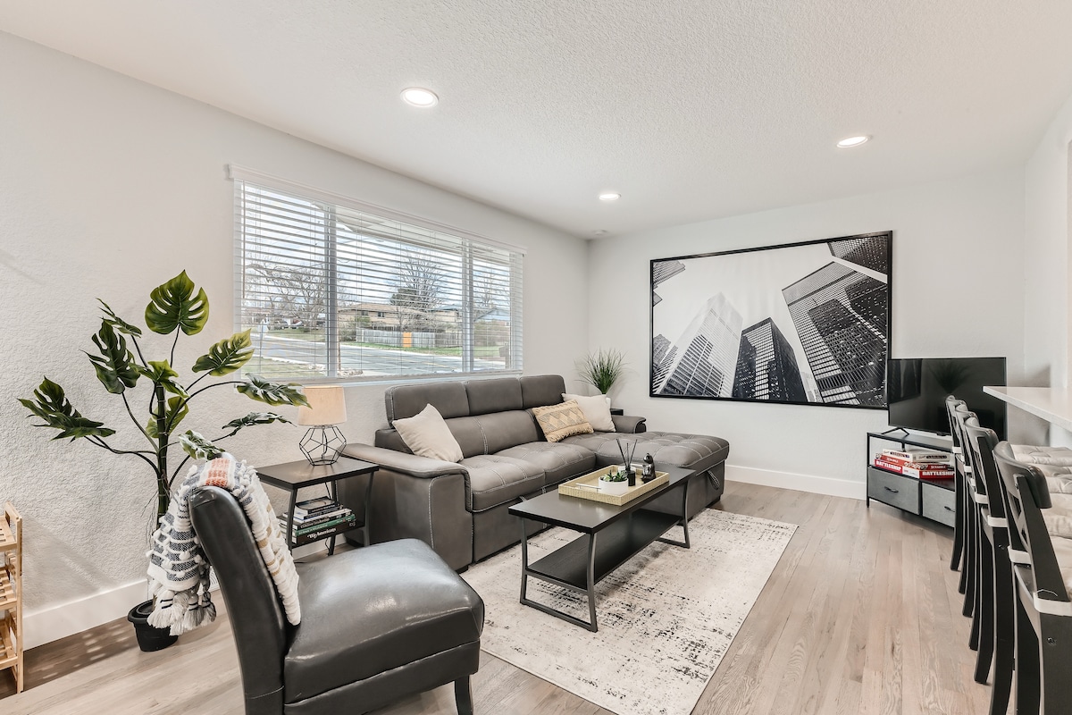 A spacious living area is highlighted by a large, framed black and white cityscape photograph. A modern sectional sofa with decorative cushions is positioned beside a sleek coffee table. There is ample natural light from windows, with greenery adding a touch of nature to the decor.
