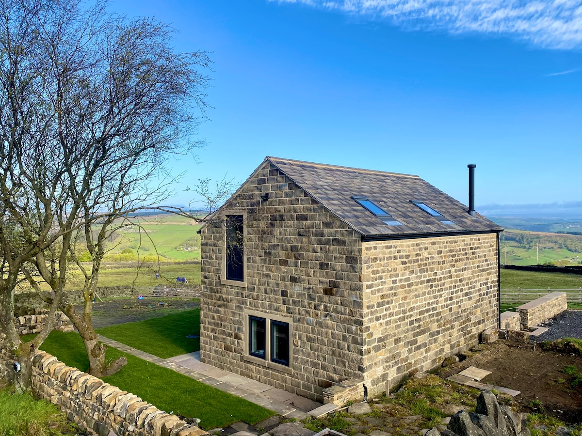 A stone cottage stands amidst expansive greenery, showcasing a modern design. Large windows are present, allowing for natural light, and the property is bordered by a low stone wall. The surrounding landscape features hills under a clear blue sky.