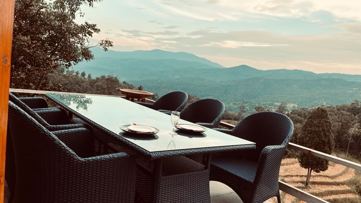 A dining area is set outdoors with a large glass table surrounded by six woven chairs, providing views of surrounding mountains and valleys. The tranquil landscape features soft green hills under a cloudy sky, creating a serene atmosphere for dining.