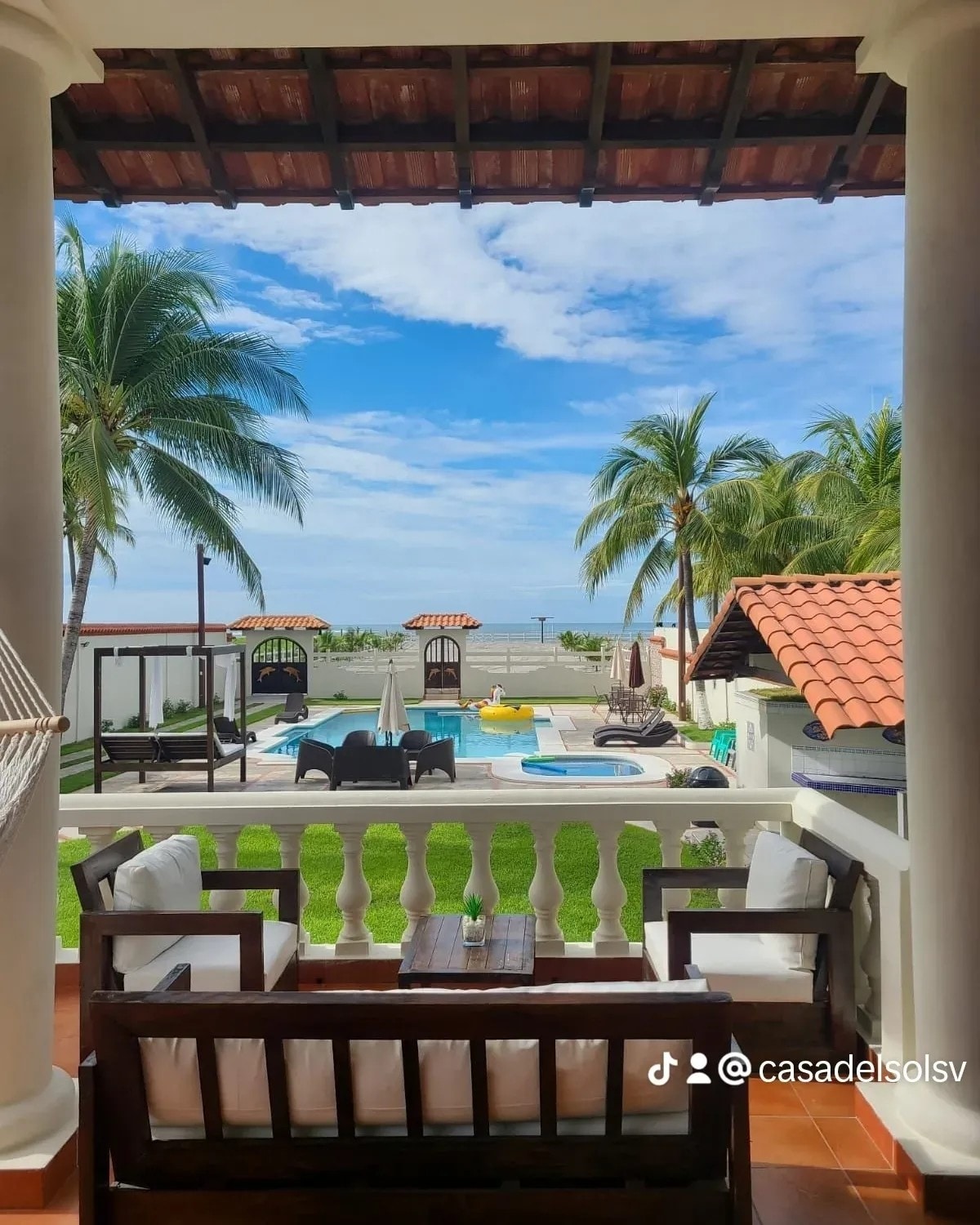 A serene view is presented from a balcony featuring comfortable seating. The outdoor area includes a large swimming pool, palm trees, and relaxation spaces with shaded structures, all overlooking the beachfront. Soft clouds are visible against a bright blue sky.