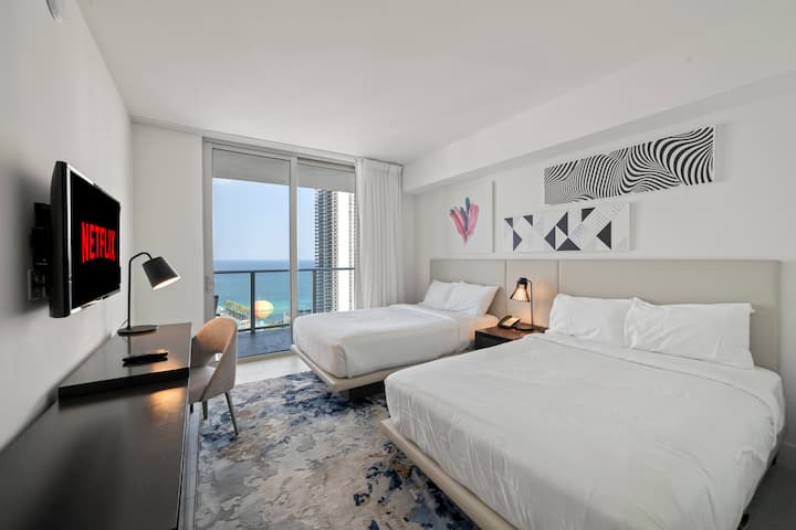 Entrez dans une deuxième chambre lumineuse avec deux lits doubles et vue sur l'océan. Un bureau et une télévision à écran plat ajoutent au confort, tandis que les lampes de chevet créent une ambiance chaleureuse. La chambre dispose également de sa propre salle de bain pour plus d'intimité et de commodité.