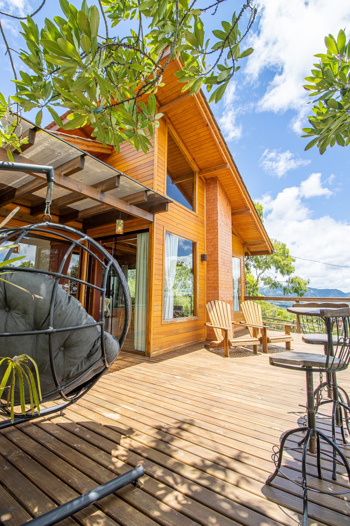 A spacious wooden deck features a hanging chair and two Adirondack chairs, surrounded by lush greenery. Large windows offer views of the outdoors, and a small table with two chairs is set for outdoor dining, inviting relaxation and enjoyment of the fresh air.