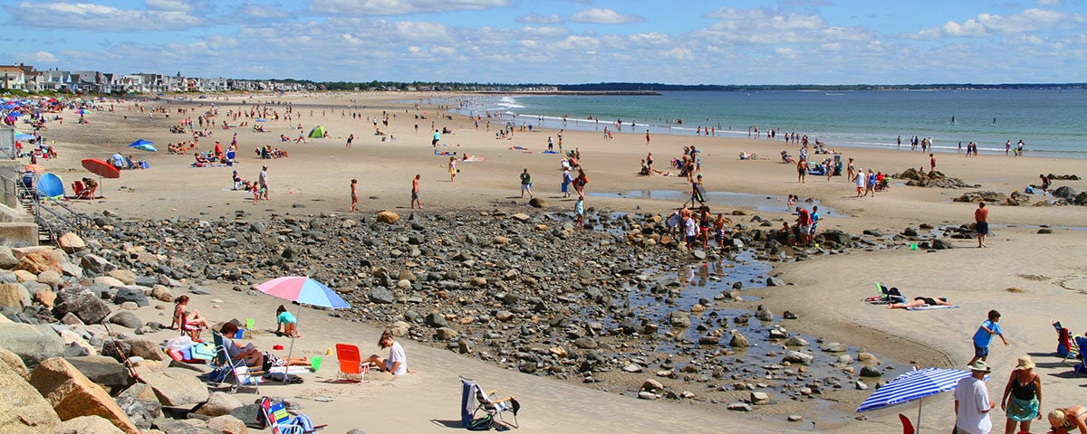A wide sandy beach is populated with visitors enjoying the day. Colorful umbrellas provide shade for families scattered along the shore. Rocky outcrops and tidal pools are visible, while the ocean stretches toward a distant horizon under a partly cloudy sky.