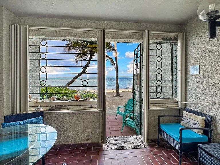 A welcoming entryway is framed by open glass doors, revealing a view of the beach and turquoise waters. Two blue chairs and a small glass table sit on a tiled floor, complimented by greenery in the foreground. Palm trees are visible in the background.