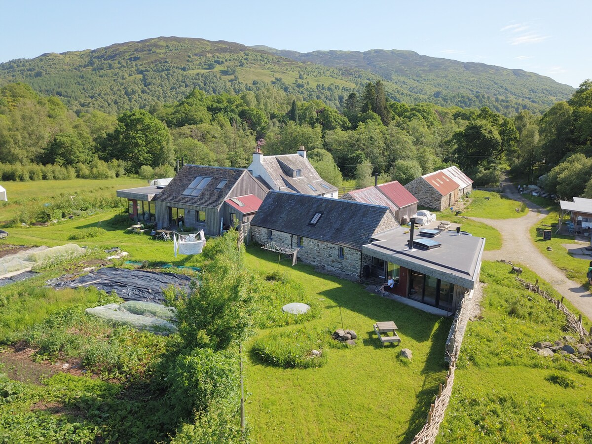 An aerial view of Achray Farm reveals a combination of traditional and modern buildings nestled within lush greenery. The farm features stone structures with varied roof colors, surrounded by cultivated land and a tree-lined backdrop, highlighting the secluded rural setting.