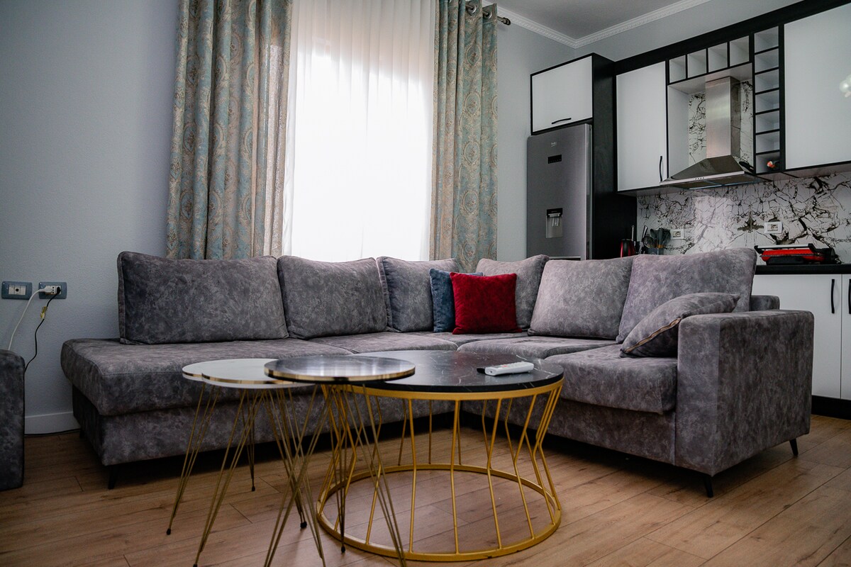 A spacious living area is presented, featuring a comfortable gray sectional sofa adorned with a red accent pillow. Two round coffee tables with gold bases are positioned in front. Natural light filters through the sheer curtains, creating a bright and inviting space.