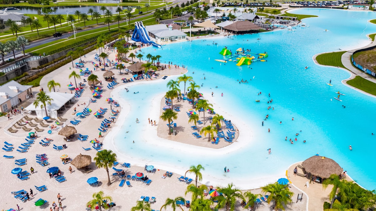 The expansive water park features vibrant blue waters, with various slides and flotation devices visible. The area includes sandy beach space with shaded seating and palm trees. Groups of people are seen enjoying the water and surrounding amenities, contributing to a lively environment.