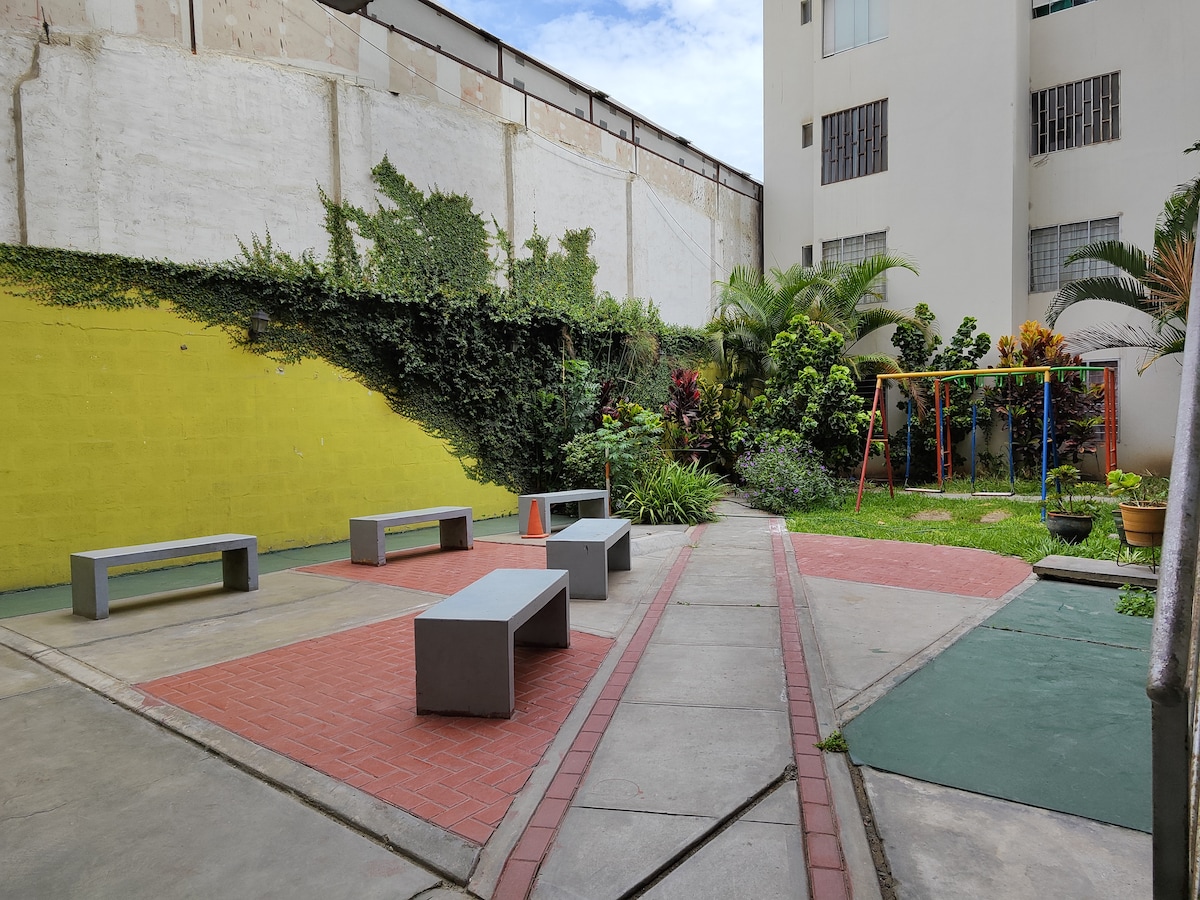 A communal outdoor area is presented, featuring concrete benches and paved walkways. Lush greenery and flowering plants are visible alongside colorful wall sections, while a swing set is situated in the background, inviting relaxation and play.