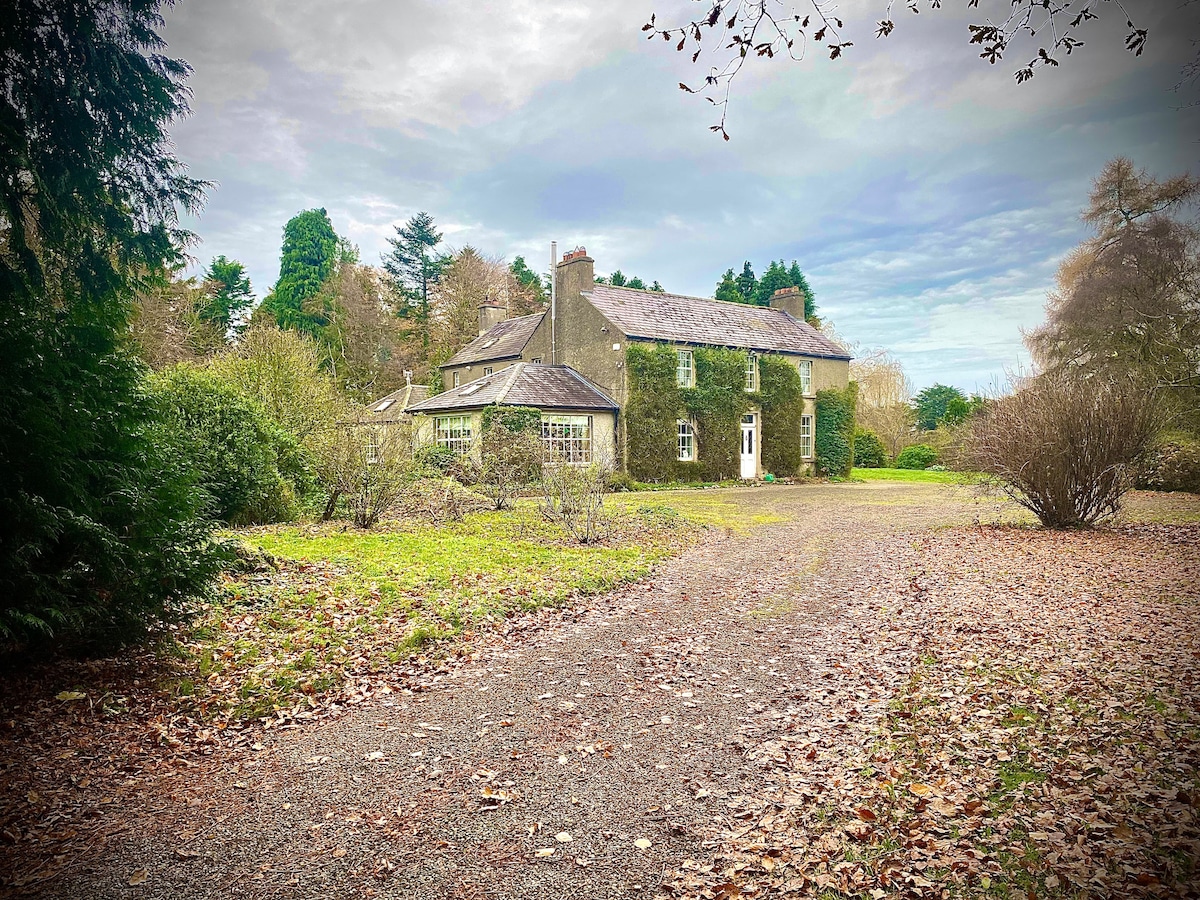 The historic 16th-century apartment is set among lush greenery, bordered by a gravel path. Scattered leaves cover the ground, while a grand stone structure, adorned with natural vines, rises from the landscape. The serene environment is complemented by the expansive garden space.