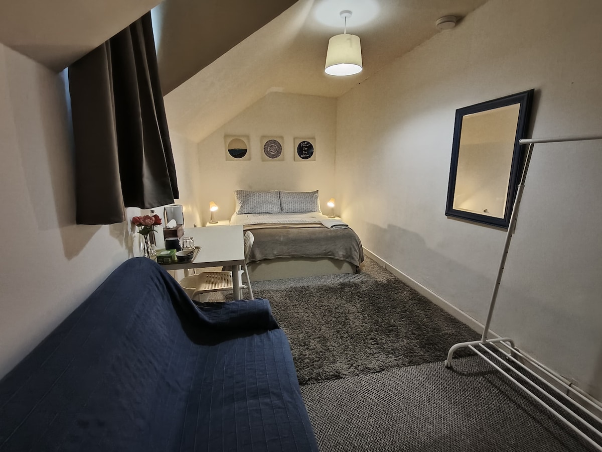 A spacious double room features a comfortable bed with a light-colored blanket. A small table with flowers is positioned beside the bed, and a full-length mirror is mounted on the wall. Soft lighting creates an inviting atmosphere in the room with textured carpet flooring.