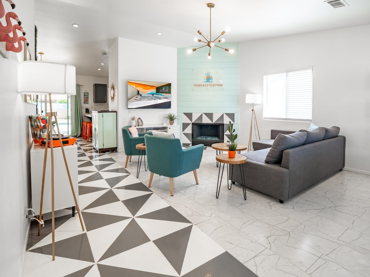 An inviting living area showcases modern furnishings, including a gray sofa and two teal armchairs positioned around a geometric-patterned rug. A flat-screen TV is mounted on the wall, while a decorative fireplace with a light blue accent wall is visible. Natural light filters through blinds on the window.