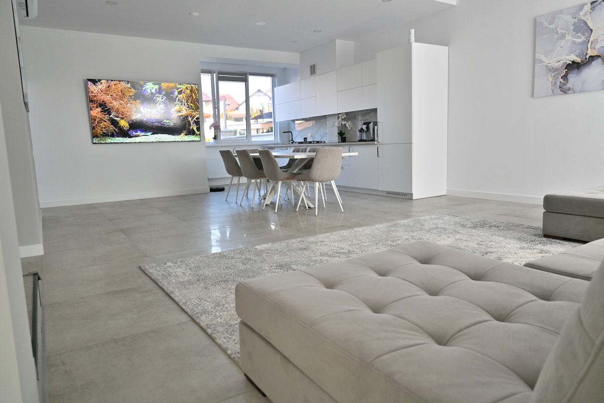 A modern living area features an open layout with a comfortable sectional sofa and a large area rug. A dining table with four chairs is positioned near the kitchen, which showcases sleek cabinetry. A wall-mounted television displays a scenic landscape.