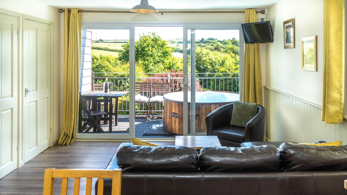 The spacious living area features large glass doors that open onto a balcony with scenic views of the surrounding countryside. A private hot tub is visible on the deck. Warm curtains frame the view, and comfortable seating arrangements are present within the room.