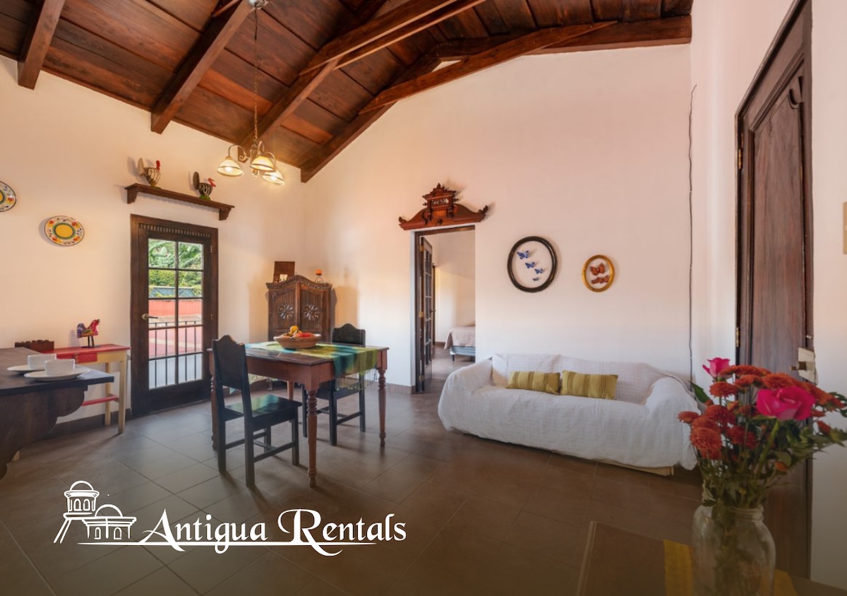 A bright living area is displayed, featuring a dining table surrounded by chairs and a cozy white sofa. Decorative plates are hung on the walls, while large windows allow natural light to fill the space. The ceiling is adorned with wooden beams.