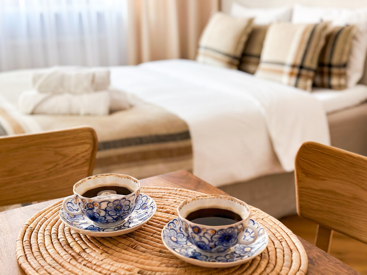 A cozy setup showcases two ornate cups of coffee on a woven placemat, positioned on a wooden table. A spacious bed with neutral-colored linens and plaid-accented pillows is visible in the background, complemented by soft, natural light filtering through a nearby window.
