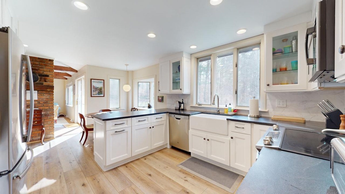 A spacious kitchen features modern cabinetry with white finishes and a contrasting dark countertop. Natural light streams through multiple windows, highlighting the stainless steel appliances and inviting work areas. A dining space is visible in the adjacent room, creating a warm atmosphere for gatherings.