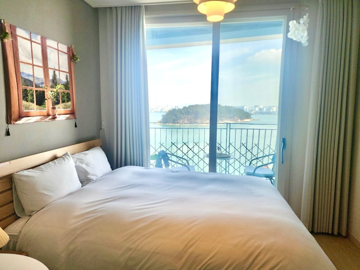 A comfortable sleeping area features a spacious bed with light bedding. A large glass door leads to a balcony with a clear ocean view, complemented by sheer curtains. A photo of a window with a scenic landscape decorates the wall beside the bed.