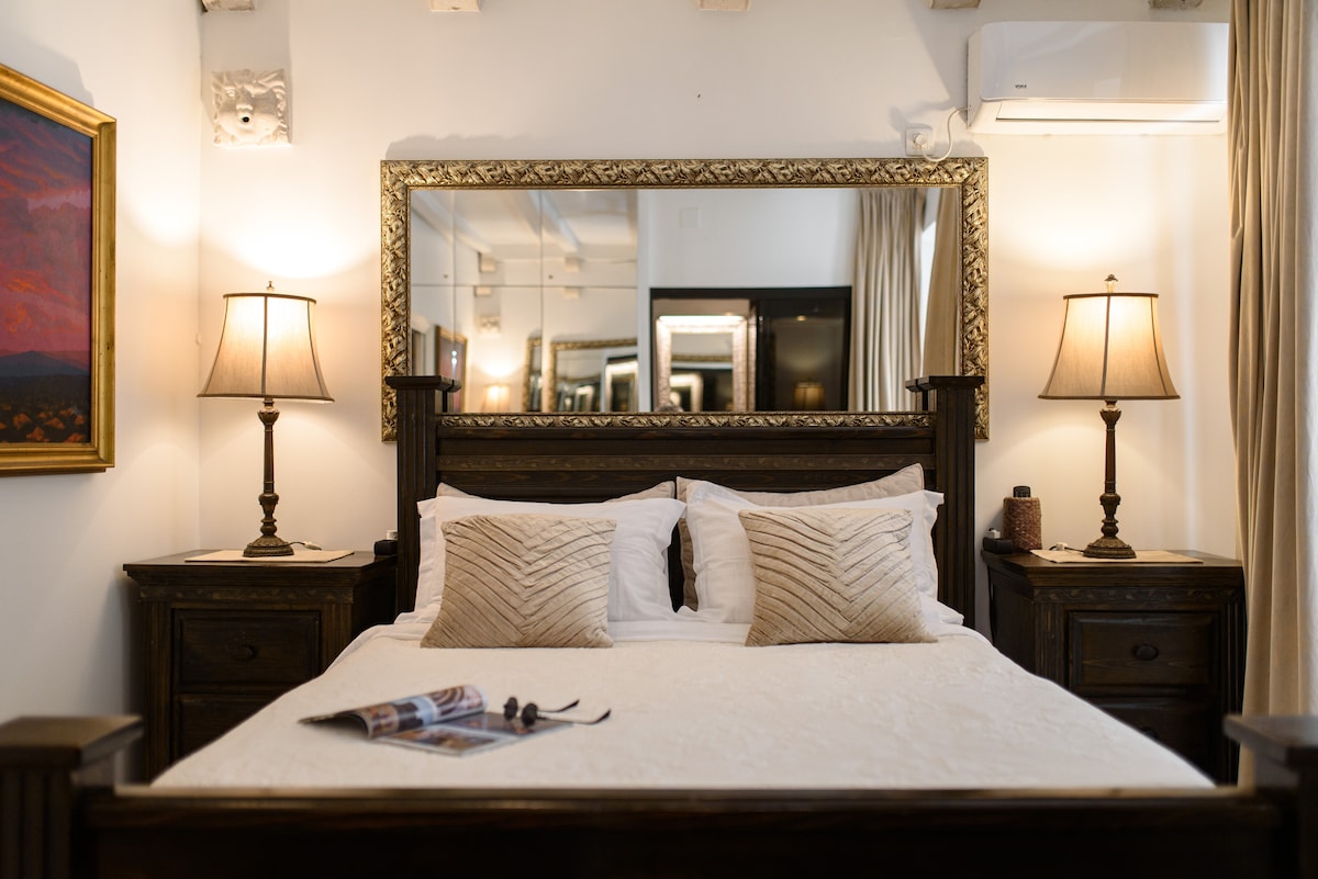 A cozy bedroom features a wooden bed frame with a white quilted comforter and decorative pillows. Matching bedside tables are positioned on either side, each with a lamp and decorative items. A large mirror is mounted above the bed, reflecting the soft lighting and elegantly adorned walls.