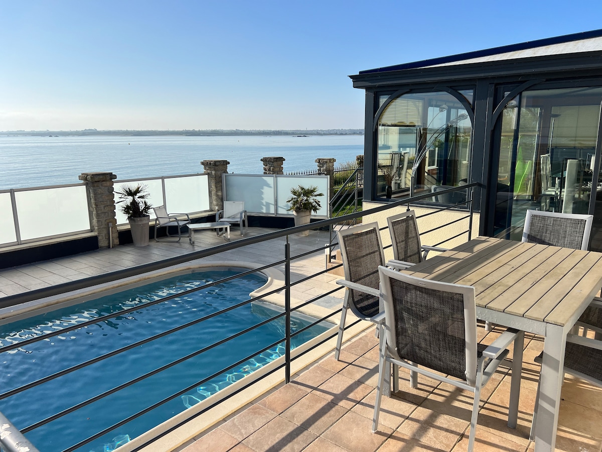 An outdoor dining area is set beside a private swimming pool, with views of the water beyond. The table is surrounded by six chairs, while a glass enclosure offers an unobstructed view of the serene landscape. Bright sunlight illuminates the scene.