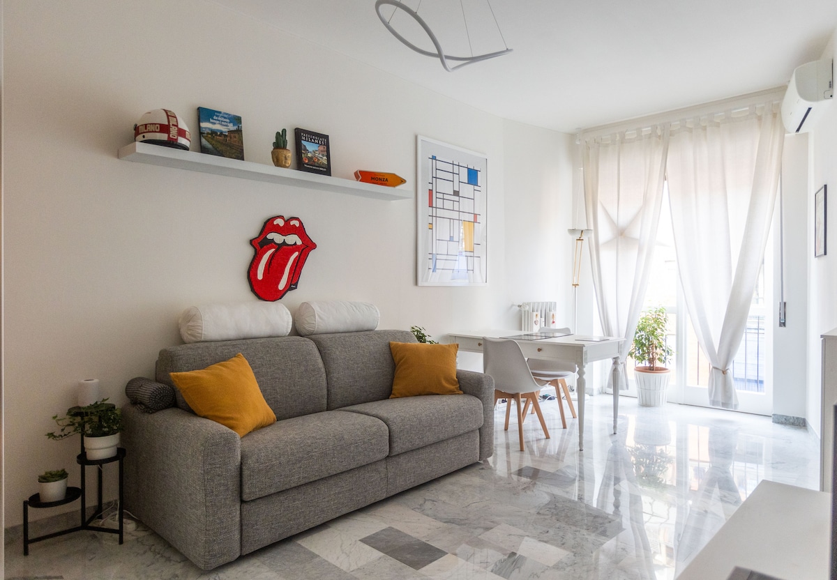 A modern living area is furnished with a gray sofa accented by yellow pillows. A stylish wall shelf displays decorative items. Natural light filters through sheer curtains, illuminating the space, while a small desk and plant add a touch of greenery.