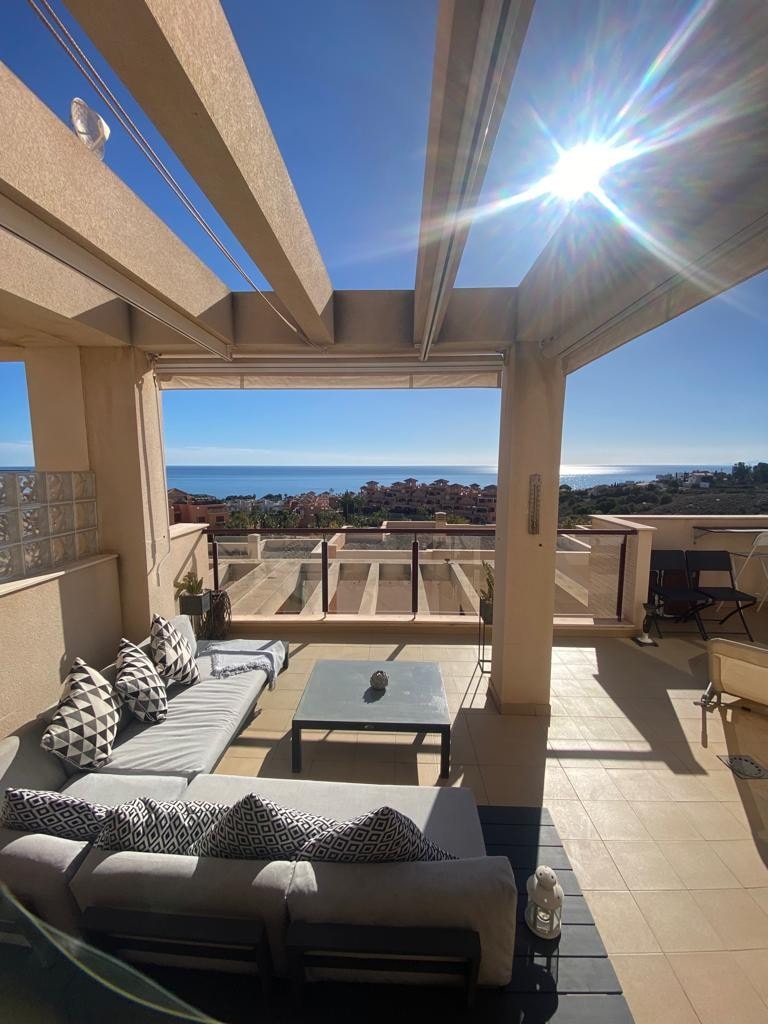 An outdoor patio is highlighted, featuring a comfortable lounge area with patterned cushions and a glass table. The space is bathed in sunlight, with a clear view of the Mediterranean Sea and surrounding landscape visible beyond the railing.