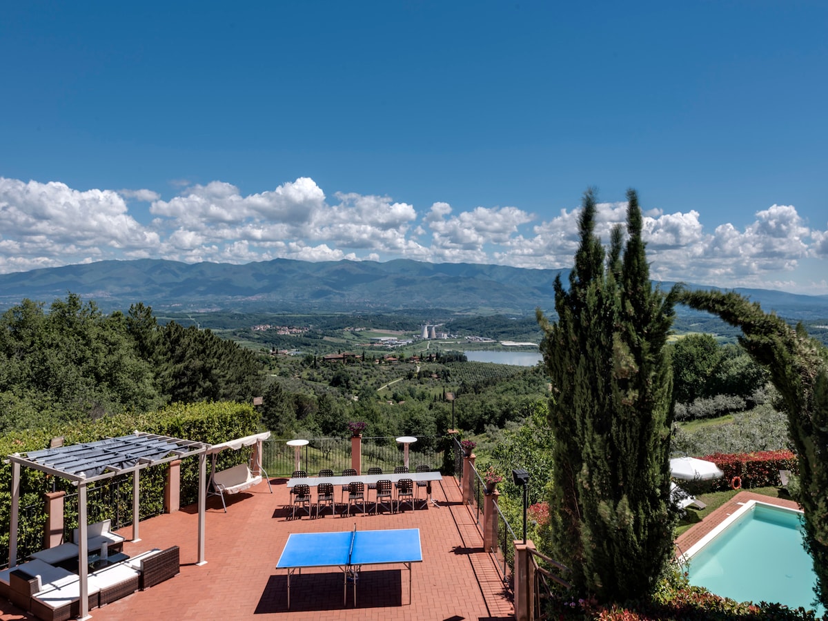 A spacious terrace features lounging chairs and a shaded seating area with a pergola. The inviting swimming pool is visible nearby, surrounded by lush greenery. Rolling hills and distant mountains create a picturesque backdrop under a clear blue sky.