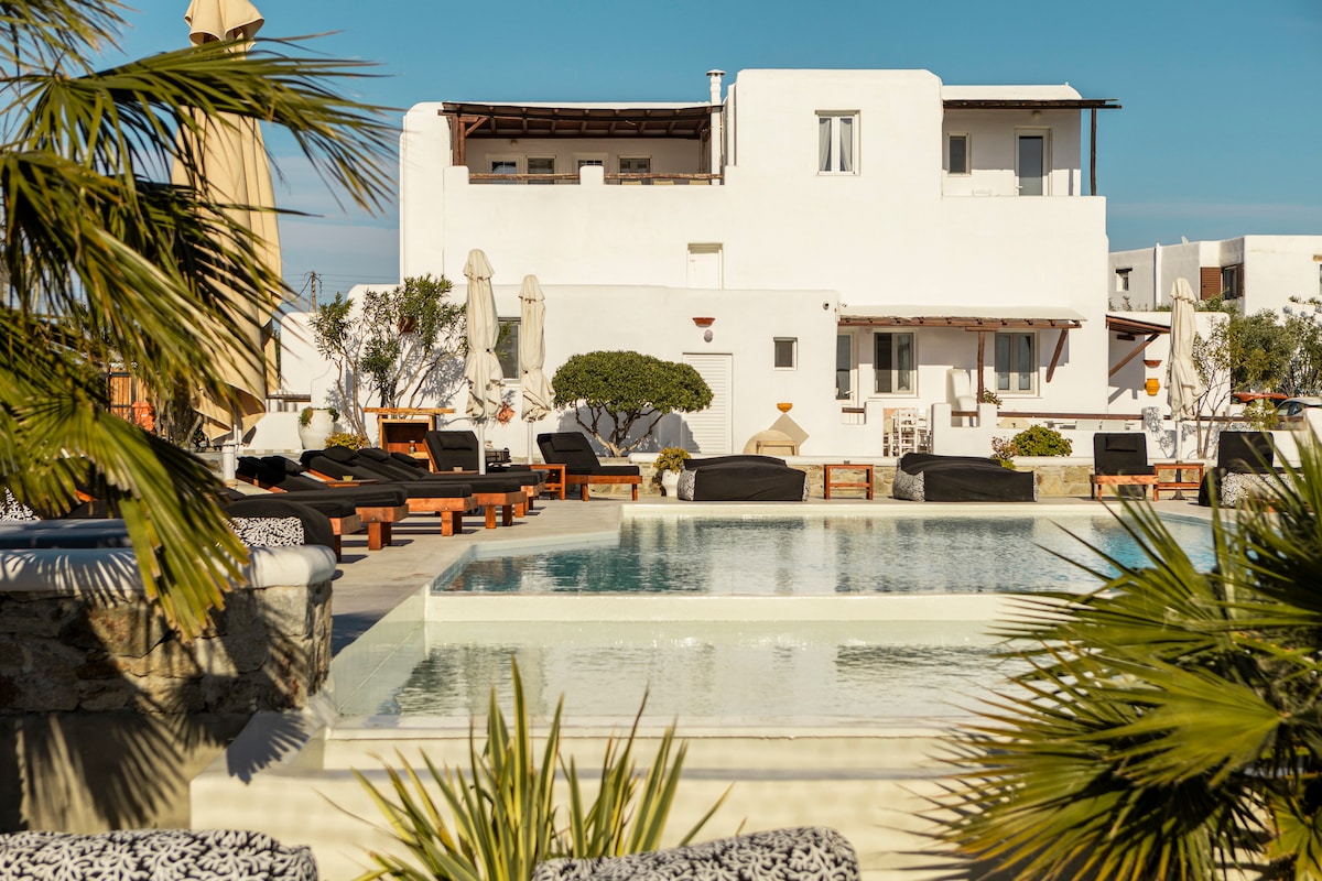 A serene pool area is surrounded by well-maintained palm trees and sun loungers. The white buildings, featuring numerous windows and shaded areas, provide a calming backdrop against the clear blue sky, offering a tranquil setting for relaxation.